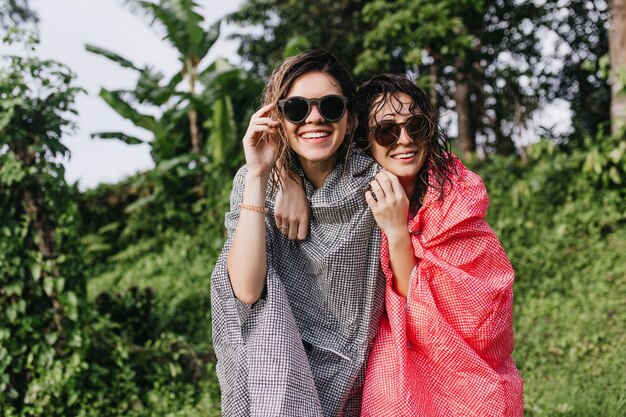 Carefree women in pink raincoat embracing sister on nature. Outdoor photo of positive ladies in sunglasses having fun in forest.