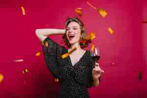 Foto gratuita giovane modella spensierata in abiti da festa che beve vino sotto coriandoli scintillanti. ridere donna bianca con taglio di capelli corto ballando all'evento.