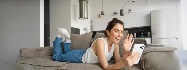 Carefree young smiling woman lying on sofa saying hello and waving at smartphone screen video