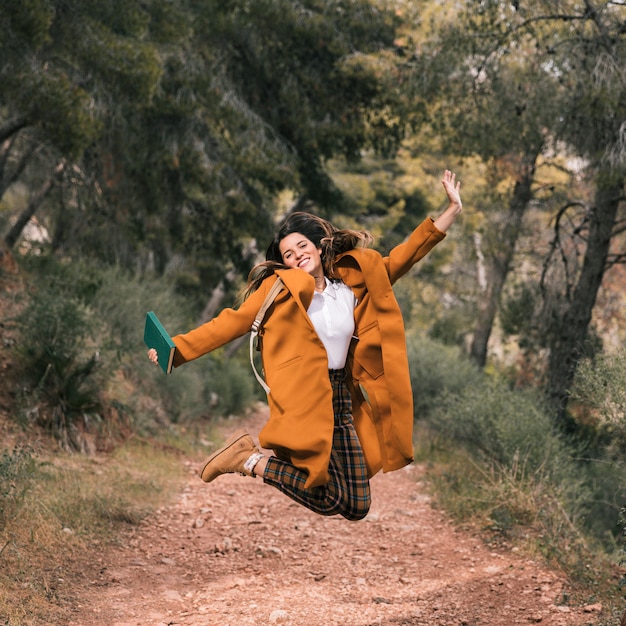 本を手で保持している屈託のない若い女性が登山道でジャンプ