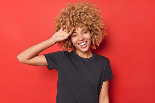 Беззаботная молодая женщина зубасто улыбается, держит руку на лбу, чувствует себя радостно, носит повседневную черную футболку и позирует на ярко-красном фоне. Позитивная кудрявая модель позирует для фото