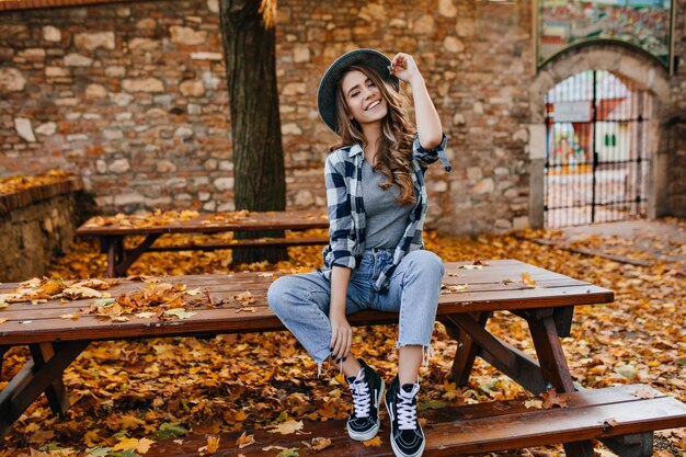 Carefree young woman in trendy vintage pants sitting on table in park and laughing