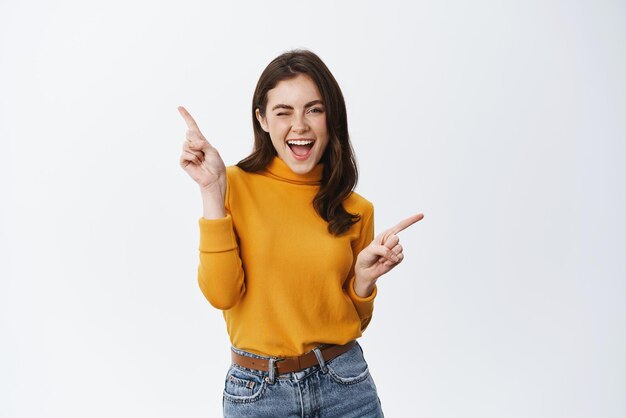 Carefree young woman winking and smiling pointing sideways at two promo offers showing advertisement standing in casual clothes against white background