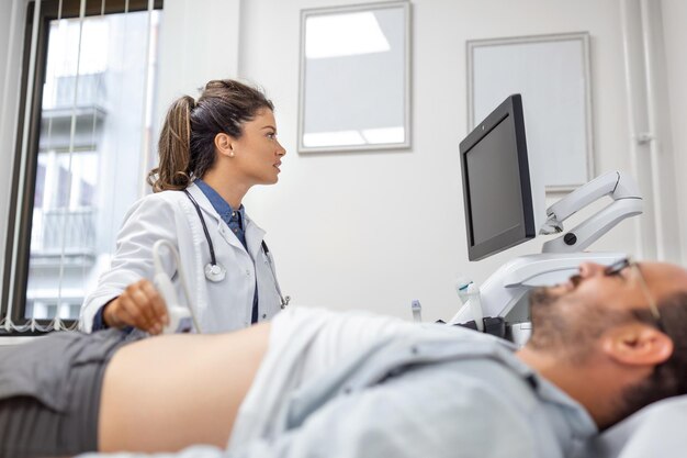 초음파 장치 앞에 앉아 변환기로 복부 진단을 수행하는 흰색 코트를 입은 신중한 여성 의사