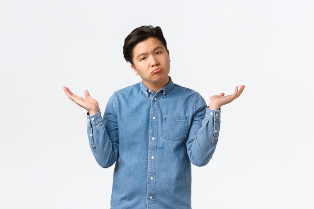 Careless asian man with no emotions, pouting as raising hands up and shrugging, dont know anything, dont care, being unaware and clueless, have no answers, standing white background.