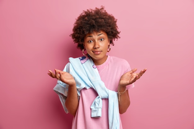 Careless indifferent woman with curly hair spreads hands sideways, has doubts and faces dilemma, cannot give answer, dressed in casual clothes, poses against pink wall, feels confused.