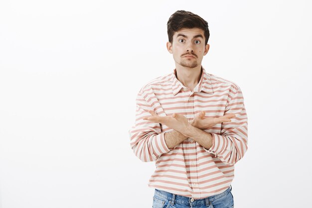 careless ordinary european male model with beard and moustache, crossing arms on chest and pointing at different directions with indifferent unaware expression, standing over gray wall