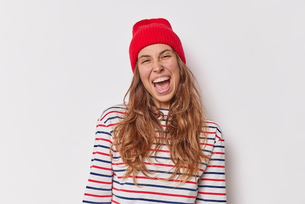 Careree positive woman laughs happily keeps mouth opened being full of joy exclaims loudly wears red hat casual striped jumper isolated over white background. People emotions and feelings concept