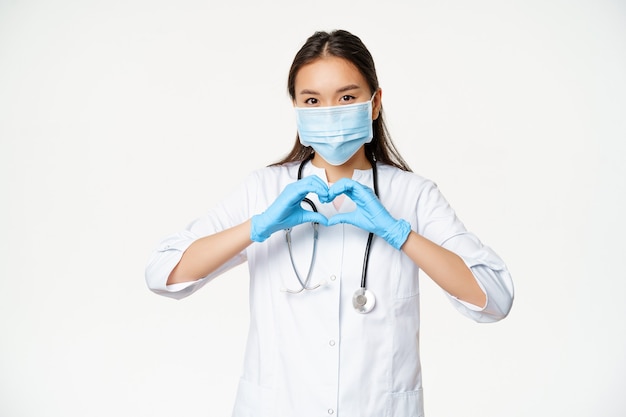 思いやりのあるアジアの医師、医療マスクとゴム手袋の女性医師は、心臓の兆候、白い背景を持つ患者の彼女のケアを示しています