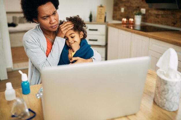 Заботливая черная мать держит свою больную дочь, используя ноутбук дома
