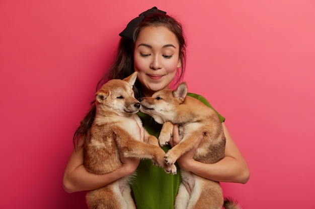 Caring compassionate female volunteer found two puppies, carries dogs to shelter, gives behavior training to pets, poses against pink background.