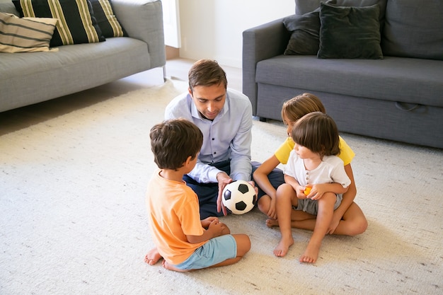 ボールを持って子供たちに話をする思いやりのお父さん。居間の床に座って一緒に遊んでいる白人の中年の父と子供たち。子供の頃、ゲーム活動と父性の概念