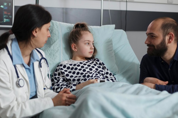 医師が治療計画を提示している間、病院の小児病棟で休んでいる病気の娘の隣に座っている思いやりのある父親。親と話し合いながら、子供の健康状態を調べる小児科医。