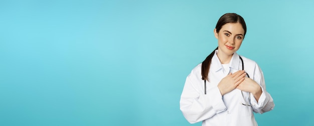 Caring healthcare worker young female doctor holding hands on heart and smiling looking with care an