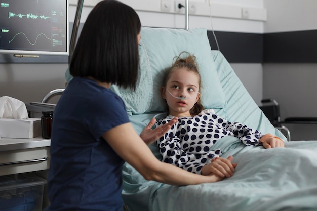 Caring parent sitting beside sick little girl while comforting her. Caring mother talking with hospitalized ill daughter resting in hospital pediatric ward patient bed while under medicine treatment.