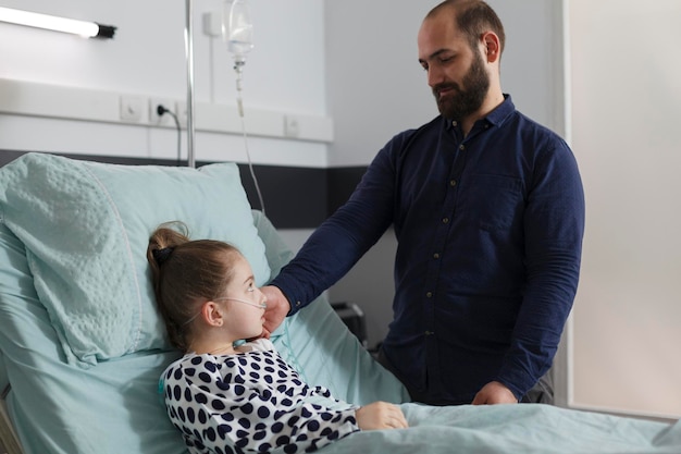 Foto gratuita genitore premuroso che si prende cura della bambina malata mentre le tocca il viso. padre amorevole che conforta la figlia malata ricoverata in ospedale che riposa nel letto del paziente all'interno della stanza del reparto pediatrico dell'ospedale