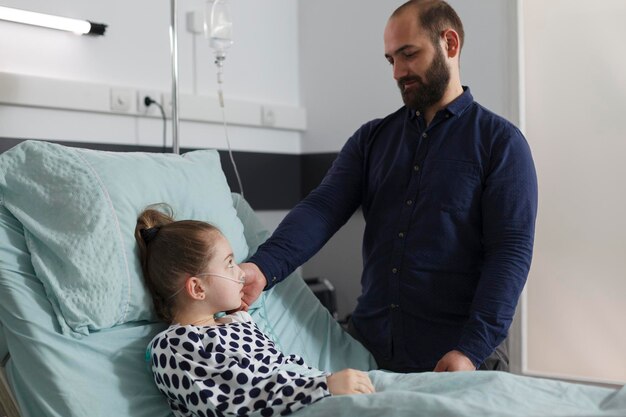 Caring parent taking care of ill little girl while touching her face. Loving father comforting hospitalized sick daughter resting in patient bed inside hospital pediatric ward room