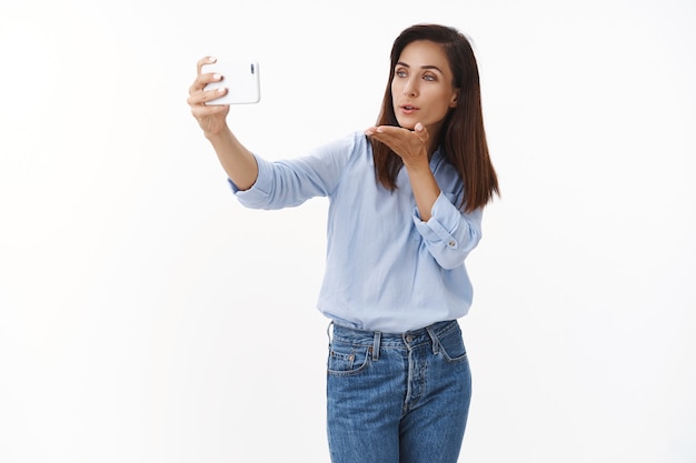 Caring tender pretty adult wife send kisses via smartphone video-call, talking partner, taking flirty selfie, blow mwah hold mobile phone, look gadget display front at front, stand white wall