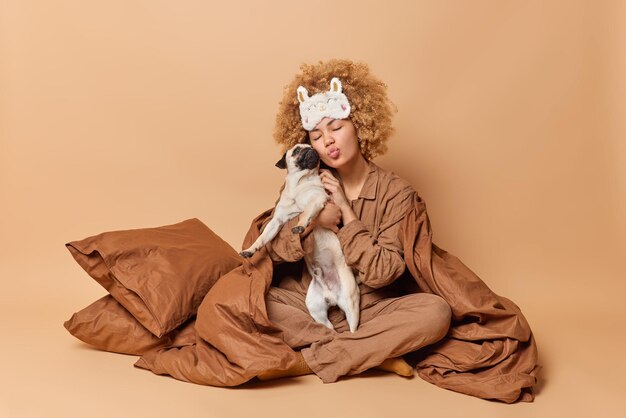 Caring woman takes care of her pet embraces pug dog with love keeps lips folded stays in bed under soft blanket enjoys time for rest isolated over brown background Domestic animals concept