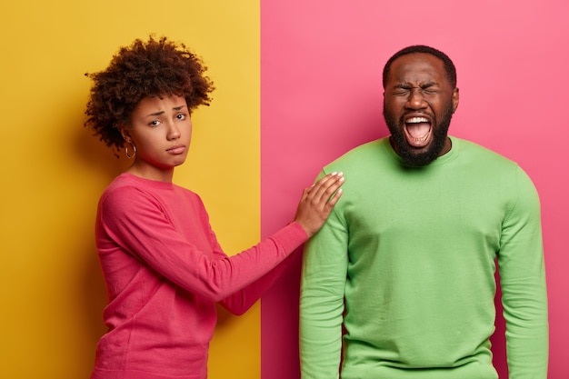 Free photo caring woman tries to calm husband, touches shoulder and persuades everything will be okay, looks with sad expression, stands sideways. depressed dark skinned man cries from despair, needs support