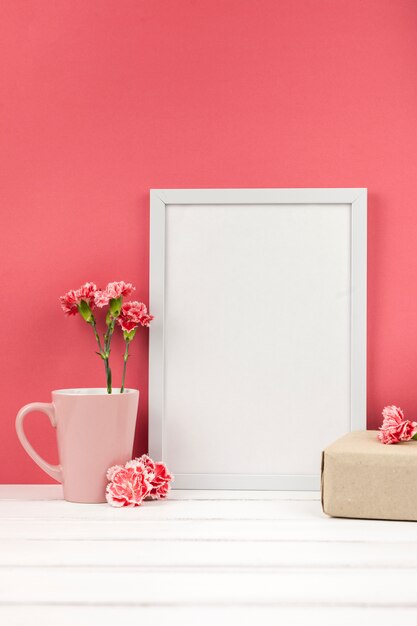 Carnation flowers; gift box; cup and white empty frame on table