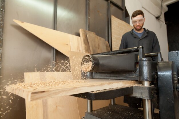 Carpenter joiner working on stationary power planer with wooden planks