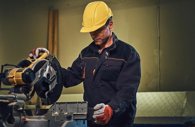 Foto gratuita il maschio del carpentiere vestito con un berretto giallo di sicurezza lavora con la motosega in un garage.