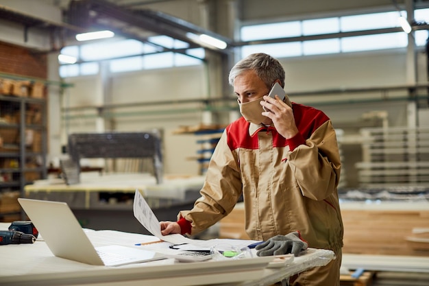 Free photo carpenter talking on the phone while using laptop and wearing face mask in a workshop