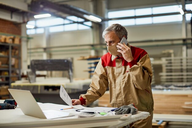 Carpenter talking on the phone while using laptop and wearing face mask in a workshop