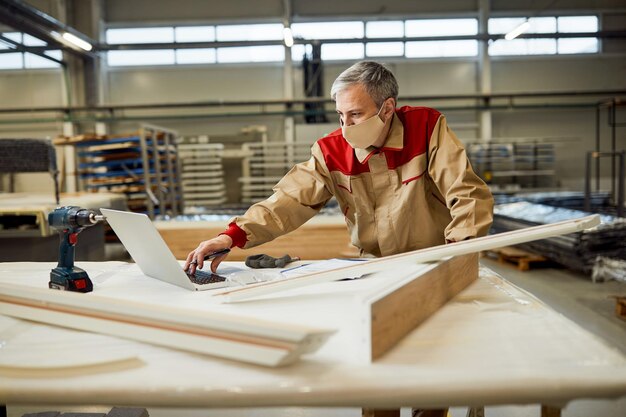 Carpenter with face mask using laptop while working in a workshop