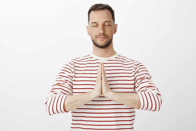 Free photo carry bad thoughts away with yoga. portrait of calm relaxed attractive guy in striped pullover, holding hands in pray and closing eyes