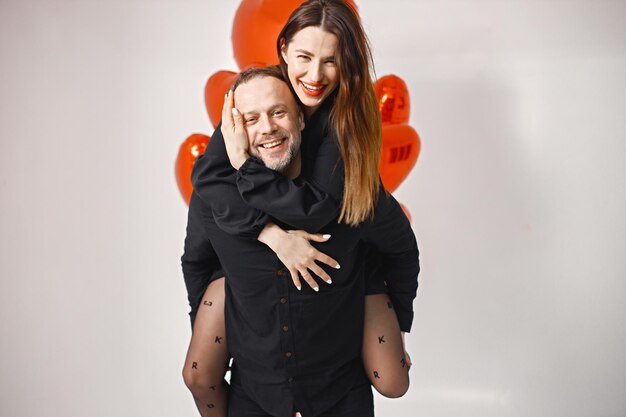 An carrying woman piggyback while posing in studio near bunch of heartshaped ballons