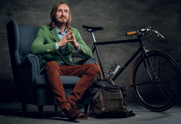 A casual bearded hipster man in a green jacket sits on a chair with single speed bicycle on background.