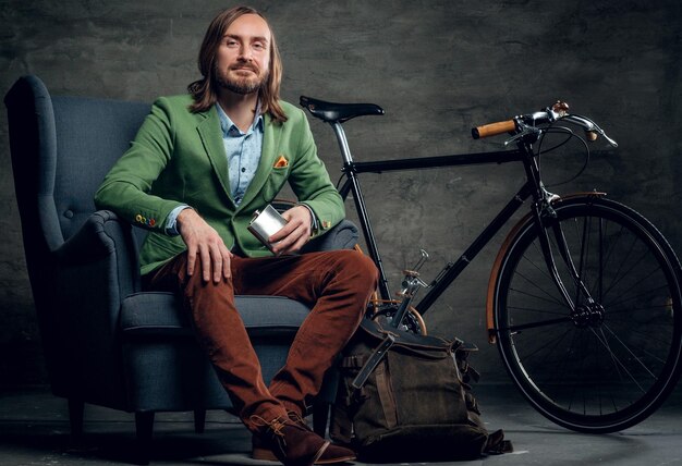 A casual bearded hipster man in a green jacket sits on a chair with single speed bicycle on background.