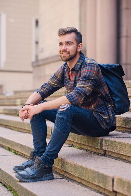 Повседневный бородатый мужчина в джинсах и флисовой рубашке сидит на ступеньке.