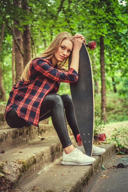 Casual blond female posing with longboard on stairs in a wild nature park.