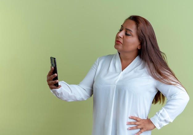 Casual caucasian middle-aged woman holding and looking at phone and putting hand on hip 