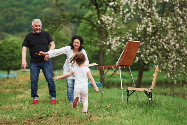 In casual clothes. Grandmother and grandfather have fun outdoors with granddaughter. Painting conception