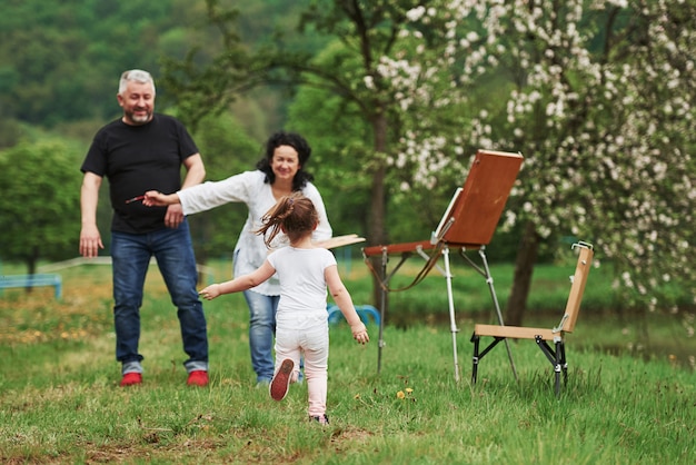 In casual clothes. Grandmother and grandfather have fun outdoors with granddaughter. Painting conception