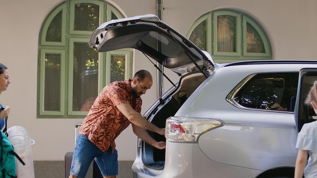 Foto gratuita famiglia informale che va in viaggio estivo mentre carica i bagagli nel bagagliaio del veicolo. madre e padre mettono bagagli e carrelli all'interno dell'auto mentre la figlia eccitata si prepara per le vacanze al mare.