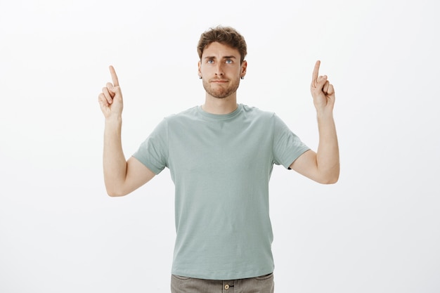 Casual good-looking male coworker in t-shirt, raising index fingers and pointing up while making indifferent annoyed grimace