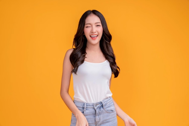 カジュアルな幸せアジアの女性女性が白いTシャツで元気に笑っているブルージーンズは、黄色の背景のスタジオ撮影で立っている平和な前向きな思考のんきなライフスタイルをリラックスします