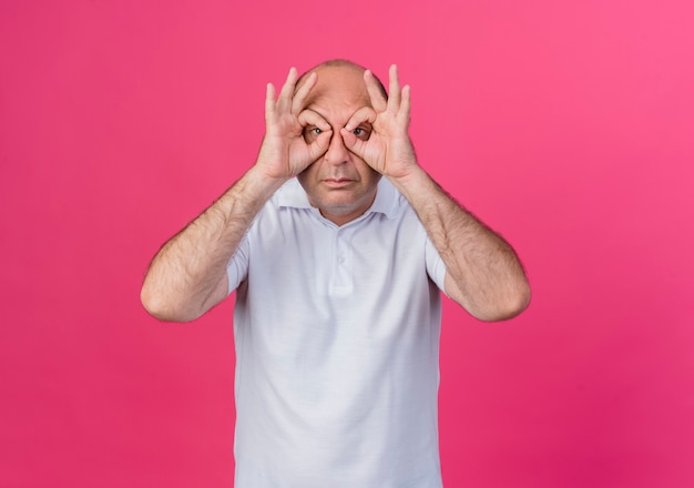 Бесплатное фото Случайный зрелый бизнесмен делает жест взгляда в камеру, используя руки как бинокль, изолированные на розовом фоне с копией пространства