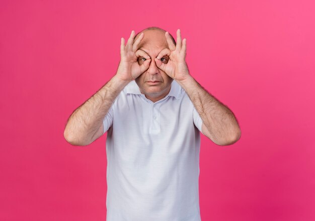 Free photo casual mature businessman doing look gesture at camera using hands as binoculars isolated on pink background with copy space