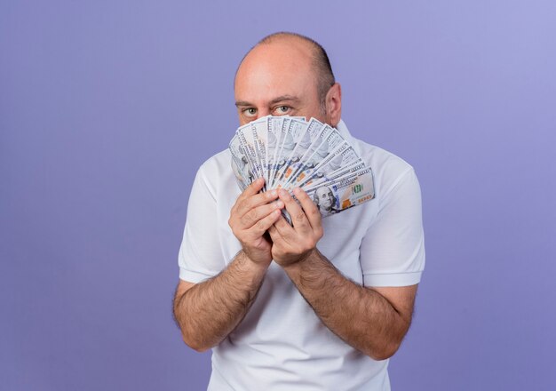 Casual mature businessman holding money and looking at camera from behind money isolated on purple background with copy space