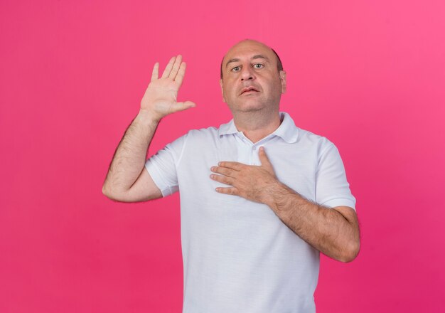 Casual mature businessman looking at camera and doing promise gesture isolated on pink background with copy space