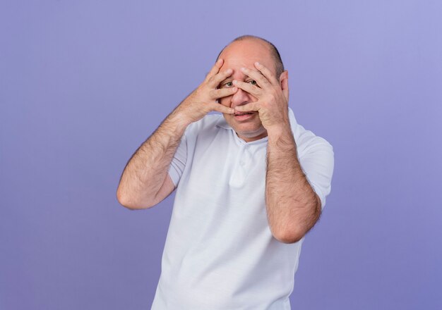 Случайный зрелый бизнесмен кладет руки на лицо и смотрит в камеру сквозь пальцы, изолированные на фиолетовом фоне с копией пространства