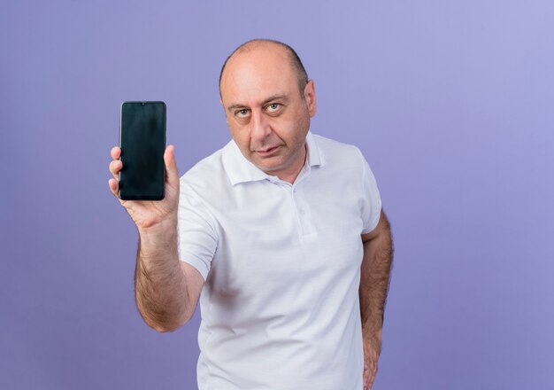 Casual mature businessman showing mobile phone and looking at camera and keeping hand on waist isolated on purple background with copy space