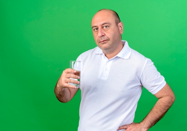  casual mature man holding glass of water and holding hand on hip isolated on green wall with copy space