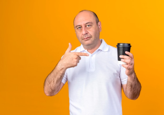casual mature man holding and points at cup of coffee isolated on yellow wall